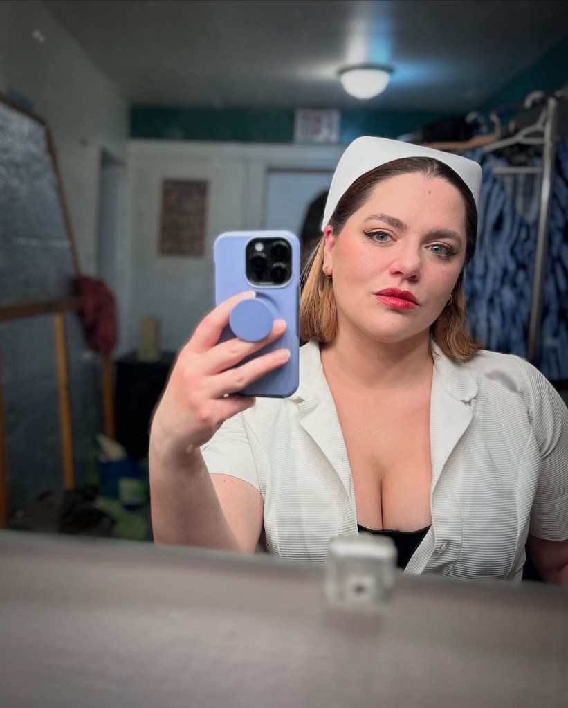 A mirror selfie of me, a white woman with shoulder-length dark hair, wearing an approximation of a vintage white nurse's uniform, the front open to showcase CLEAVAGE and a peak of a black bodysuit. I'm wearing heavy black eyeliner and red overdrawn lipstick...and a shit-ton of other makeup, but not the kind that stands out