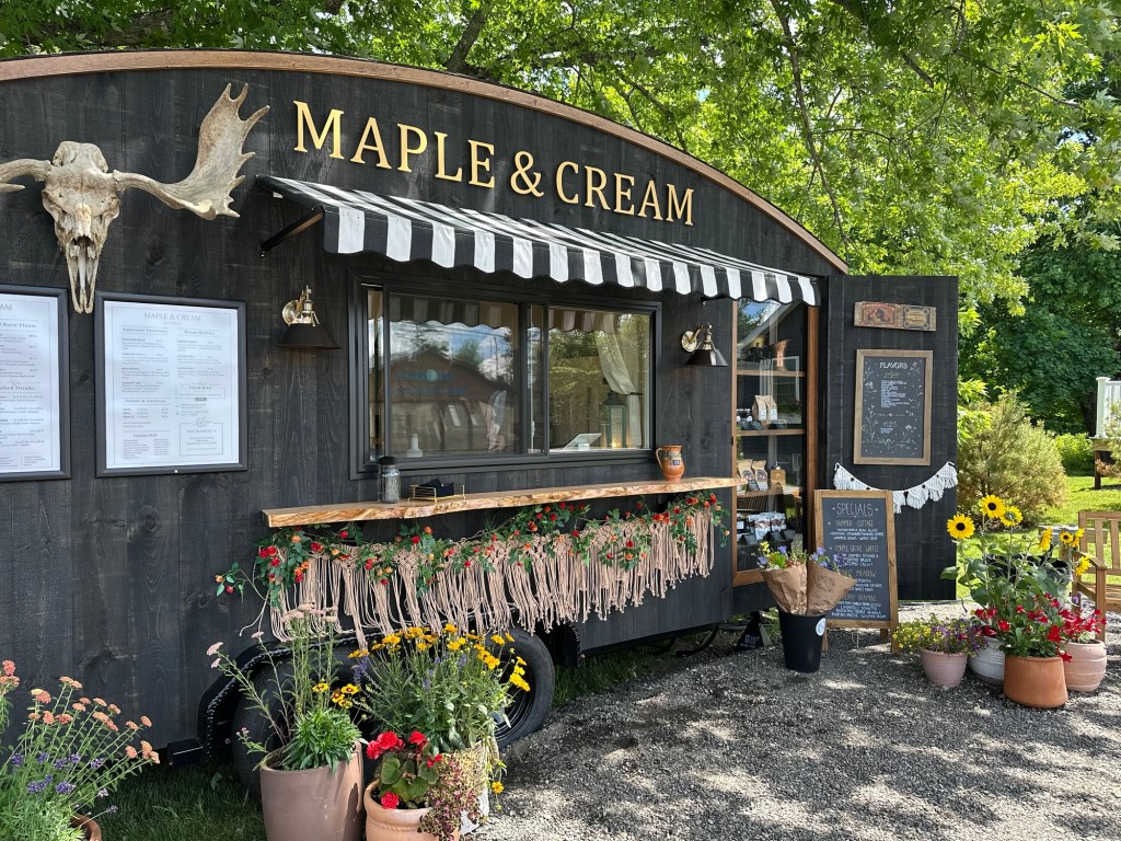 A coffee/waffles/gelato food truck (trailer) named Maple & Cream seemingly made for Instagram. It is painted black with natural wood accents and flowers in pots and hanging from the counter.